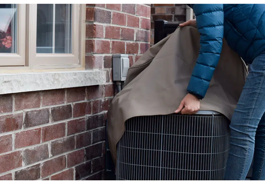  Person in blue winter jacket putting a cover over outdoor AC unit next to a brick home. 